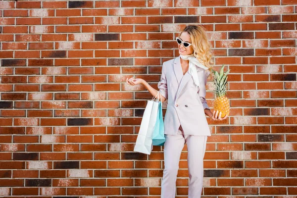 Bela jovem com abacaxi e sacos de compras em pé na frente da parede de tijolo — Fotografia de Stock