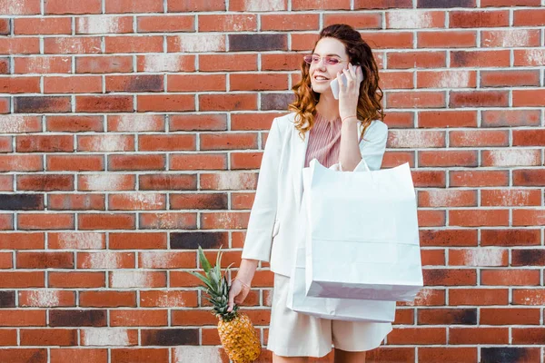 Belle jeune femme avec ananas et sacs à provisions parlant par téléphone devant un mur de briques — Photo de stock