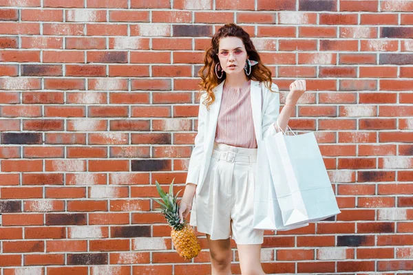 Belle jeune femme avec ananas et sacs à provisions en regardant la caméra devant le mur de briques — Photo de stock