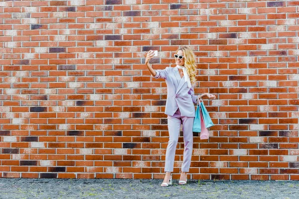 Bela jovem com sacos de compras tirando selfie na frente da parede de tijolo — Fotografia de Stock