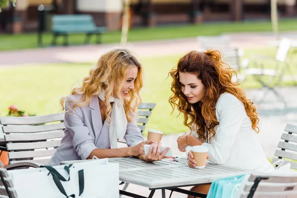 Mulheres jovens atraentes usando smartphone enquanto passam tempo juntos no café depois de fazer compras — Fotografia de Stock