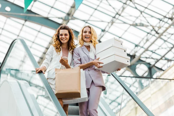 Jeunes femmes à la mode avec des sacs à provisions et des boîtes sur l'escalator au centre commercial — Photo de stock