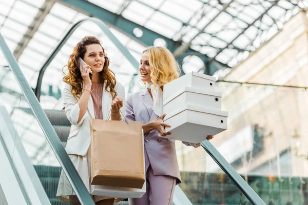 Hermosas mujeres jóvenes con bolsas de compras y cajas en escaleras mecánicas en el centro comercial - foto de stock
