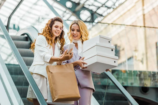 Красивые молодые женщины с сумками и коробками, глядя на смартфон на эскалаторе в торговом центре — стоковое фото