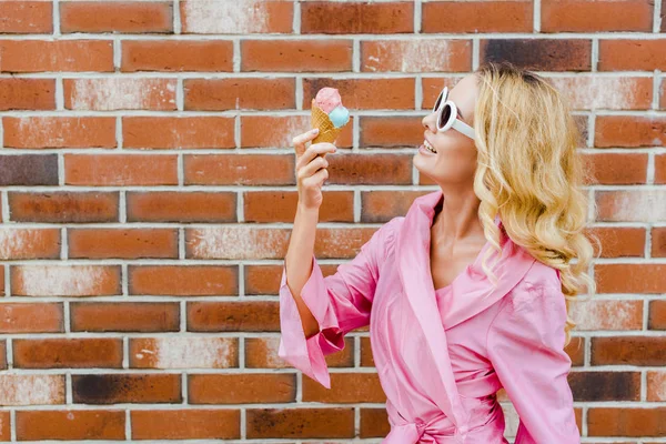Mulher sorridente na moda em rosa comendo sorvete na frente da parede de tijolo — Fotografia de Stock