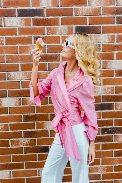 Elegante mujer sonriente en rosa comer helado en frente de la pared de ladrillo - foto de stock
