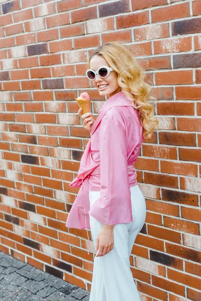 Jeune femme souriante en rose tenant la crème glacée et regardant la caméra devant le mur de briques — Photo de stock