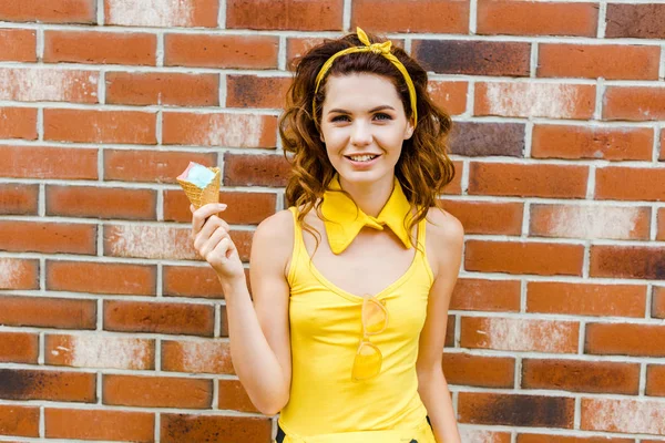 Feliz mujer joven en ropa amarilla con helado mirando a la cámara delante de la pared de ladrillo - foto de stock