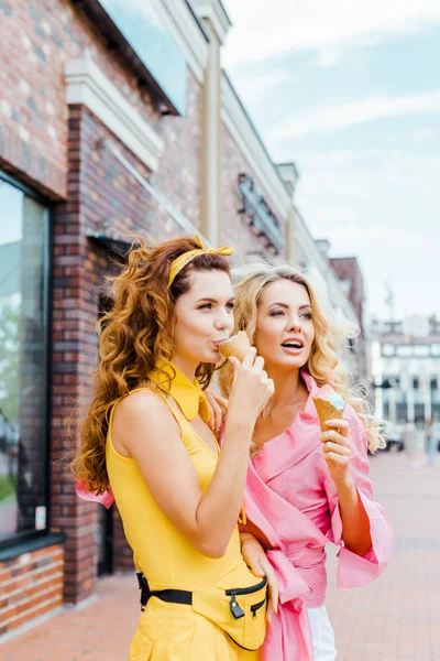 Hermosas mujeres jóvenes en ropa colorida comiendo helado en conos de gofre en la calle - foto de stock