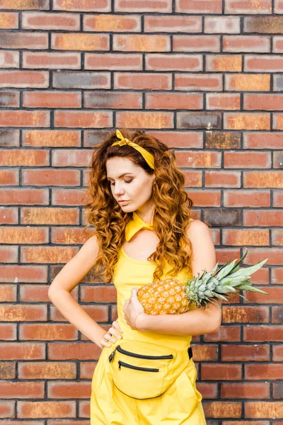 Beautiful young woman in yellow clothes holding pineapple in front of brick wall — Stock Photo