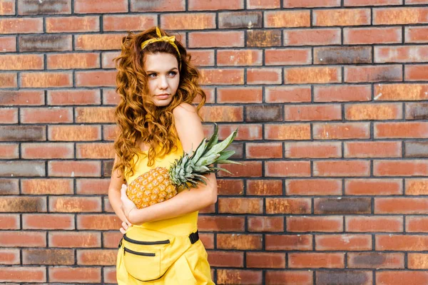 Attractive young woman in yellow clothes holding pineapple in front of brick wall and looking away — Stock Photo