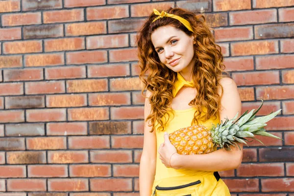 Attractive young woman in yellow clothes holding pineapple in front of brick wall and looking at camera — Stock Photo
