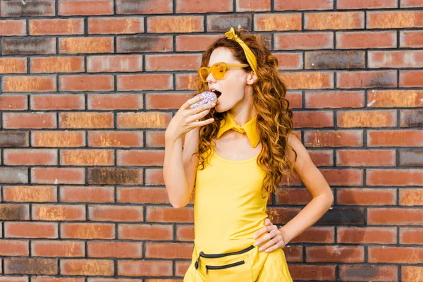 Привлекательная молодая женщина в жёлтой одежде ест пончик перед кирпичной стеной — стоковое фото