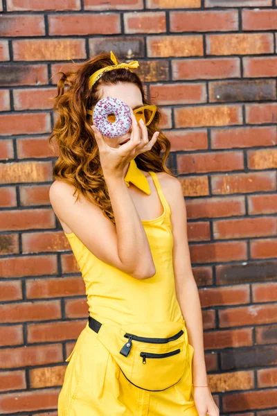 Atractiva mujer joven en ropa amarilla mirando a la cámara a través de la rosquilla delante de la pared de ladrillo — Stock Photo