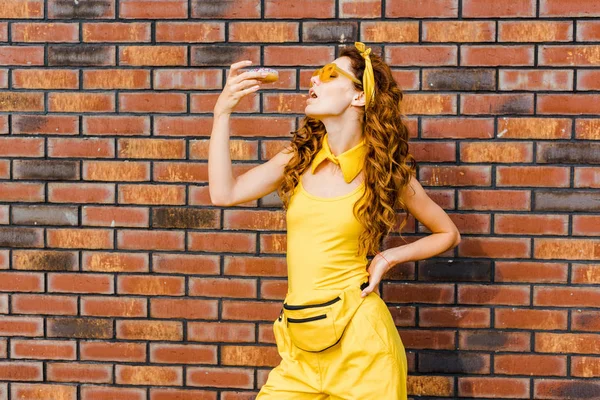 Atraente jovem mulher comer donut na frente da parede de tijolo — Fotografia de Stock