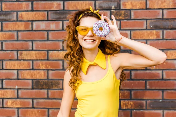 Smiling young woman in yellow clothes looking at camera through donut in front of brick wall — Stock Photo