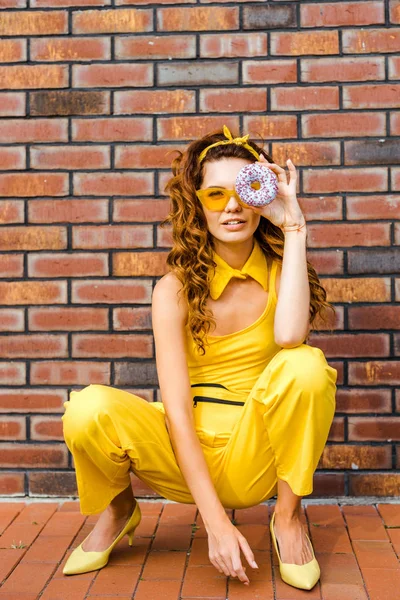 Beautiful young woman in yellow clothes looking at camera through donut sitting in front of brick wall — Stock Photo