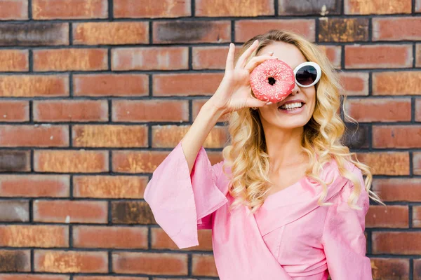 Feliz jovem mulher em camisa rosa olhando para a câmera através de donut na frente da parede de tijolo — Fotografia de Stock