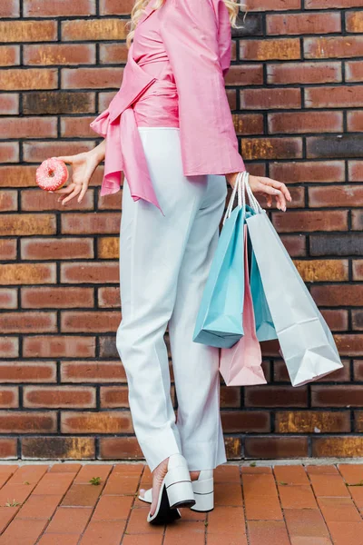 Tiro cortado de mulher com sacos de compras e donut de pé na frente da parede de tijolo — Fotografia de Stock