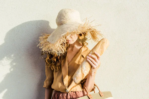 Приваблива молода жінка в стильному солом'яному капелюсі з багетом, що стоїть перед білою стіною — стокове фото
