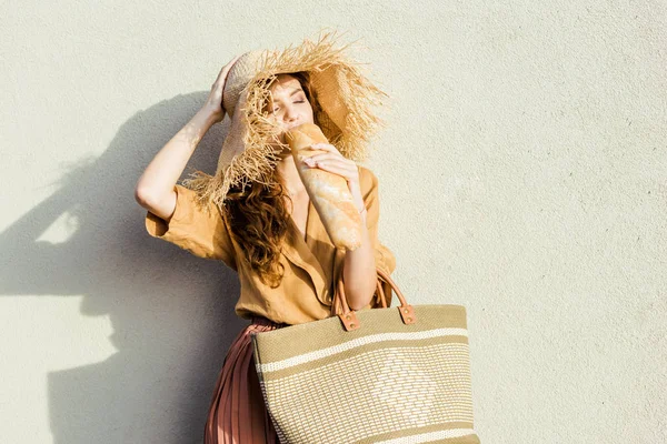 Attractive young woman in straw hat standing in front of white wall an eating baguette — Stock Photo