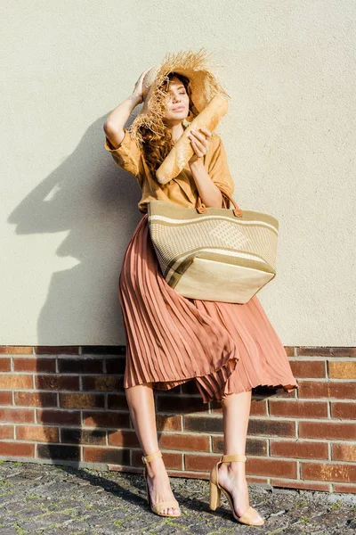 Hermosa mujer joven en sombrero de paja con baguette de pie delante de la pared blanca - foto de stock