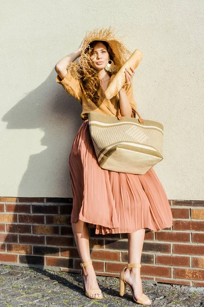 Belle femme élégante en chapeau de paille debout devant le mur blanc et manger baguette française — Photo de stock