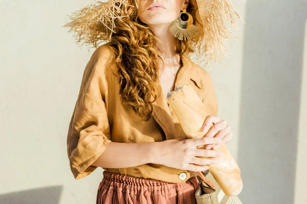 Tiro recortado de la mujer joven de moda en sombrero de paja con baguette de pie delante de la pared blanca - foto de stock