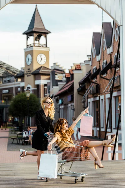 Mujeres jóvenes de moda montando carrito de compras durante las compras juntos - foto de stock