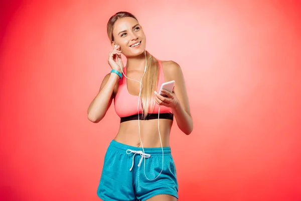 Приваблива усміхнена спортсменка, що підключає навушники та тримає смартфон ізольовано на червоному — стокове фото