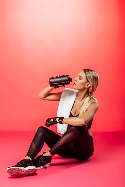 Attraktive Sportlerin sitzt mit Handtuch und trinkt Proteinergänzung aus Sportflasche auf Rot — Stockfoto