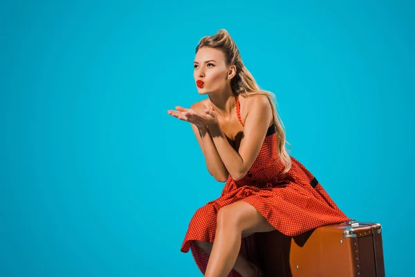 Attractive young woman in pin up style dress blowing kiss while sitting on suitcase on blue backdrop — Stock Photo