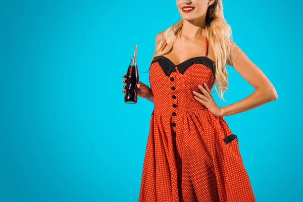 Tiro recortado de mujer sonriente en vestido vintage con bebida de soda en botella de vidrio sobre fondo azul - foto de stock