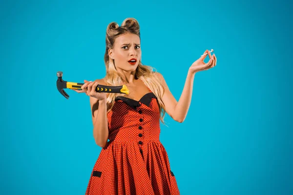 Portrait of confused pin up woman in retro dress with hammer and nail looking at camera on blue background — Stock Photo