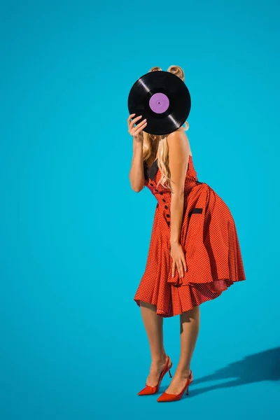 Vue obscurcie de la femme en robe vintage couvrant la tête avec disque de vinyle sur fond bleu — Photo de stock