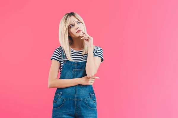 Pensive irritated girl in denim overall looking up isolated on pink — Stock Photo