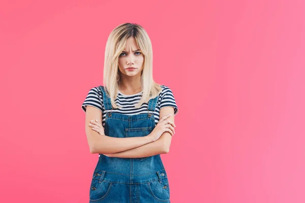 Irritated girl in denim overall standing with crossed arms and looking at camera isolated on pink — Stock Photo
