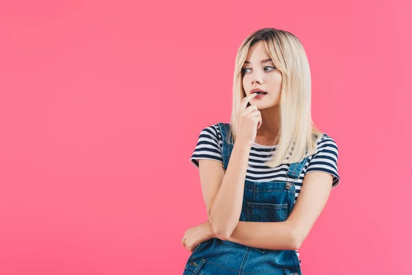 Nachdenklich schönes Mädchen in Jeans insgesamt anrührende Lippen und wegschauen isoliert auf rosa — Stockfoto