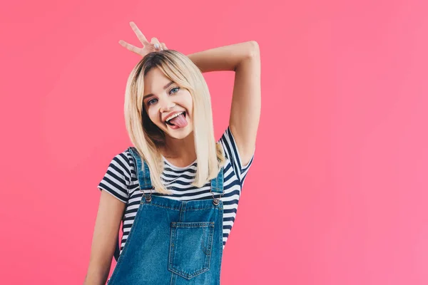 Sorridente bella ragazza in denim generale attaccare lingua fuori e mostrando orecchie di coniglio isolato su rosa — Foto stock