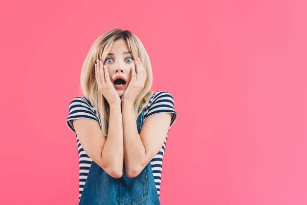 Shocked girl in denim overall touching face and looking at camera isolated on pink — Stock Photo