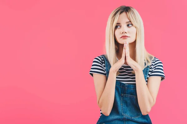 Sad beautiful girl in denim overall looking away with hands in namaste gesture isolated on pink — Stock Photo