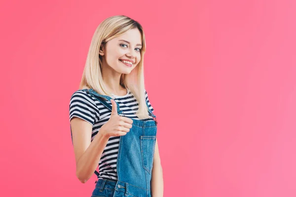 Lächeln schönes Mädchen in Jeans Overall zeigt Daumen nach oben isoliert auf rosa — Stockfoto