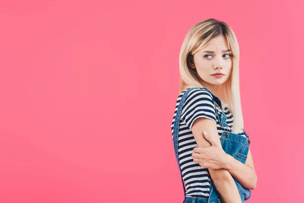 Sad girl in denim overall hugging herself and looking away isolated on pink — Stock Photo