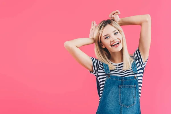 Happy beautiful girl in denim overall showing bunny ears isolated on pink — Stock Photo