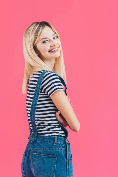 Smiling beautiful girl in denim overall standing with crossed arms and looking at camera isolated on pink — Stock Photo