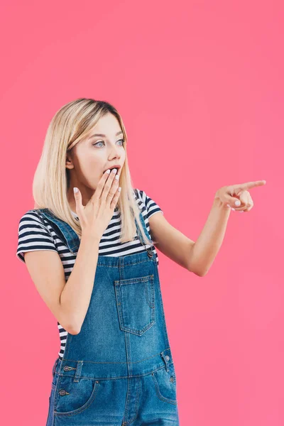 Menina chocada em jeans geral apontando em algo e cobrindo a boca isolada em rosa — Fotografia de Stock