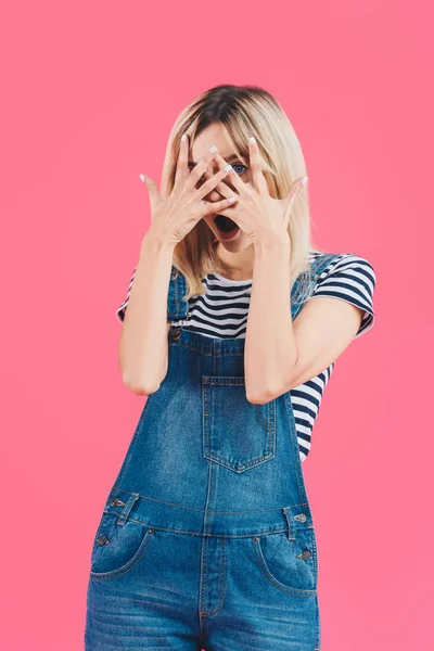 Schöne schockierte Mädchen in Jeans Overall Blick in die Kamera durch Finger isoliert auf rosa — Stockfoto