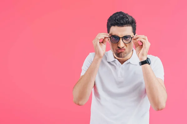 Hombre guapo haciendo muecas, tocando gafas y mirando a la cámara aislada en rosa - foto de stock