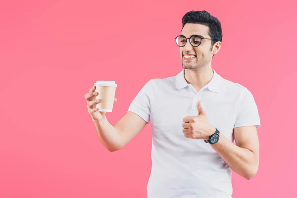 Homem bonito feliz olhando para o café em copo de papel e mostrando polegar para cima isolado em rosa — Fotografia de Stock