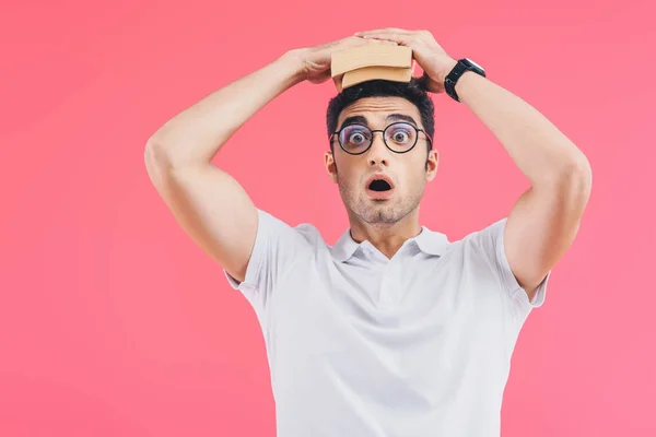 Bello studente scioccato tenendo libri sulla testa e guardando la fotocamera isolata su rosa — Foto stock